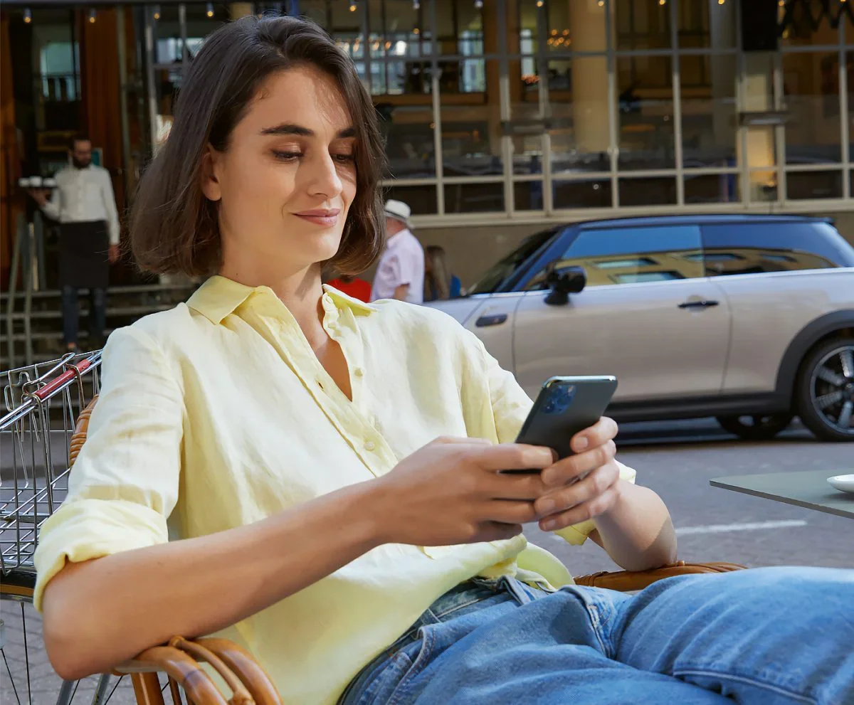 Female wearing yellow collared button-down shirt and jeans, sitting in a chair and holding a smartphone next to a street with a grey MINI vehicle parked in the background. | Ferman MINI of Tampa Bay in Palm Harbor FL