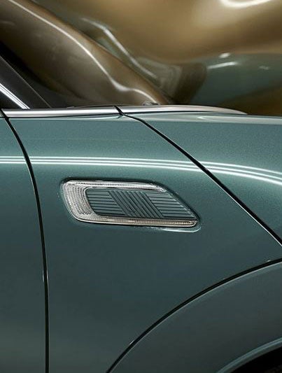 Closeup of the Sage Green metallic paint with piano black exterior trim on a MINI Clubman Untold Edition with a giant CGI, inflating, shiny, brass balloon coming from the side of the car